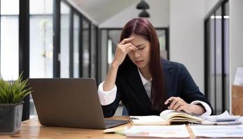 mujer asiática joven que siente tensión en la cabeza con migraña. mujer de negocios cansada y sobrecargada de trabajo mientras trabajaba en una computadora portátil en la oficina. foto