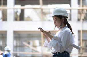 Architect with a blueprints at a construction site. Portrait of woman constructor wearing white helmet and safety yellow vest. Upset sad, skeptical, serious woman looking at the phone screen outdoors. photo