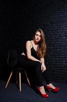 una guapa morena vestida con tacones altos negros y rojos, sentada y posando en una silla en el estudio contra una pared de ladrillo oscuro. retrato de modelo de estudio. foto