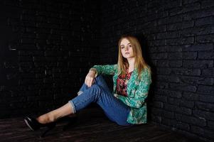 Stylish blonde girl in jacket and jeans against brick black wall at studio. photo