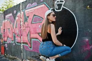 Back of buttocks stylish casual hipster girl in cap, sunglasses and jeans wear, listening music from headphones of mobile phone against large graffiti wall with large tnt bomb. photo