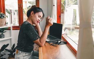 hermosa mujer asiática se sienta en el mostrador del bar en una cafetería con una taza de café y sonríe relajadamente después de haber terminado de trabajar en línea en su computadora portátil. foto