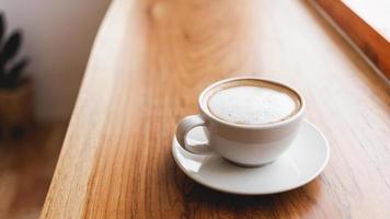 taza de café capuchino con un café con leche en una barra de madera en un café iluminado por el sol por la mañana. foto