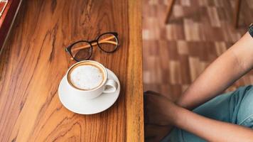 Cup of cappuccino coffee with latte and glasses of a woman on a wooden bar counter in a sunlit cafe in the morning. photo