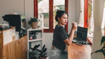 hermosa mujer asiática se sienta en el mostrador del bar en una cafetería con una taza de café y sonríe relajadamente después de haber terminado de trabajar en línea en su computadora portátil. foto