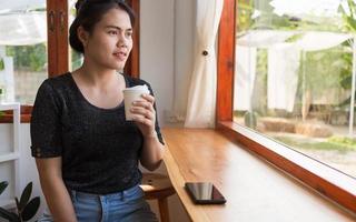 A beautiful Asian woman sits down at the bar counter inside a window coffee shop holding a paper coffee cup, smiling relaxed in a cafe photo