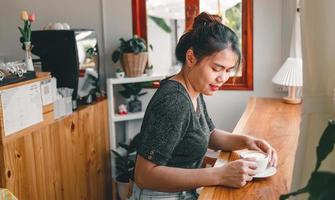 hermosa mujer asiática se sienta en el mostrador del bar dentro de una cafetería con una taza de café, sonriendo relajada en un café foto