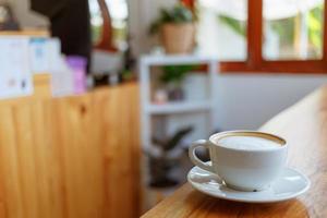 taza de café capuchino con un café con leche en una barra de madera en un café iluminado por el sol por la mañana. foto