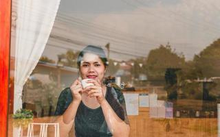 Portrait beautiful Asian woman sits down at the bar counter in a coffee shop holding a coffee cup viewed through glass with reflections as she looks at camera and smiling relaxed in a cafe photo