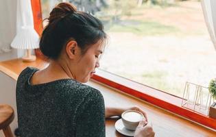 Beautiful Asian woman sits down at the bar counter inside a window coffee shop holding a cup of coffee, smiling relaxed in a cafe photo