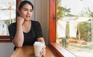 A beautiful Asian woman sits down at the bar counter inside a window coffee shop holding a paper coffee cup, smiling relaxed in a cafe photo