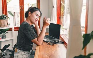 hermosa mujer asiática se sienta en el mostrador del bar en una cafetería con una taza de café y sonríe relajadamente después de haber terminado de trabajar en línea en su computadora portátil. foto