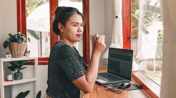 hermosa mujer asiática se sienta en el mostrador del bar en una cafetería con una taza de café y sonríe relajadamente después de haber terminado de trabajar en línea en su computadora portátil. foto