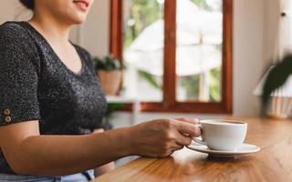 hermosa mujer asiática se sienta en el mostrador del bar dentro de una cafetería con una taza de café, sonriendo relajada en un café foto