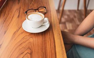 taza de café capuchino con café con leche y vasos de una mujer en un mostrador de madera en un café iluminado por el sol por la mañana. foto