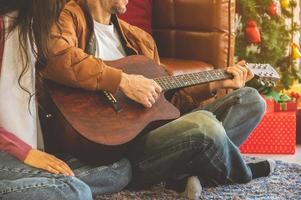 feliz pareja alegre disfrutando de un gran momento en la sala de estar con guitarra en las vacaciones de navidad. hombre tocando la guitarra y cantando para la mujer en el piso de la alfombra foto