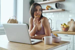 hermosa joven usando una laptop y comiendo galletas mientras se sienta en el mostrador de la cocina foto