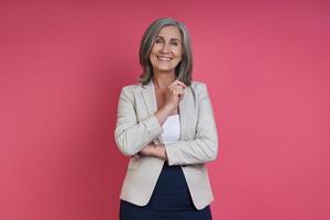 Confident senior woman in formalwear smiling while standing against pink background photo