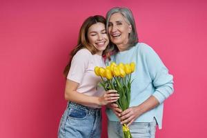 Happy senior mother and adult daughter holding a bunch of tulips against pink background photo