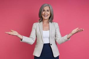 Happy senior woman in formalwear gesturing while standing against pink background photo