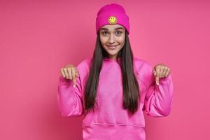 Beautiful young woman in funky hat pointing down and smiling against pink background photo