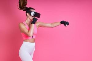 Concentrated young woman in virtual reality glasses boxing against pink background photo