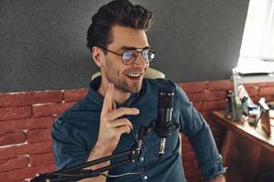 joven feliz usando micrófono y gesticulando mientras graba podcast en el estudio foto