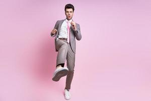 Furious young man in full suit throwing leg kick while standing against pink background photo