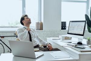 Tired young man adjusting his necktie while sitting at his working place in office photo