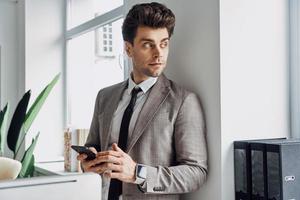 Handsome young man holding smart phone and looking through a window in office photo