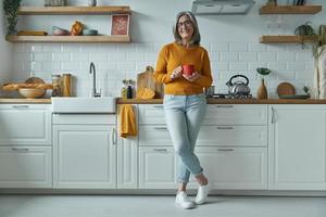 Cheerful senior woman holding cup and smiling while standing at the domestic kitchen photo