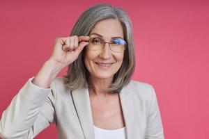 Cheerful senior woman in formalwear adjusting eyeglasses while standing against pink background photo