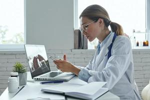 Confident female doctor using smart phone while sitting at the medical office photo