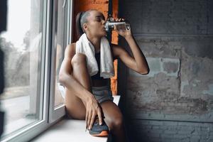 Attractive young African woman drinking water while sitting on the window sill in gym photo