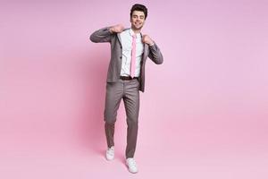 un joven alegre con traje completo ajustando su chaqueta mientras se enfrenta a un fondo rosa foto