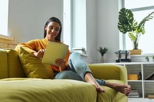 mujer joven feliz usando tableta digital mientras se sienta en el sofá en casa foto