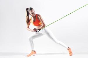 Confident young woman using resistance band while exercising against white background photo