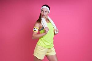 Confident woman in sports clothing carrying towel on shoulders against pink background photo