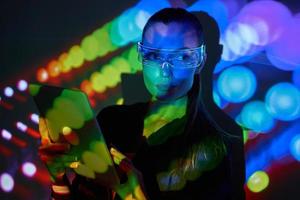 Beautiful young woman in futuristic glasses using digital tablet against colorful background photo