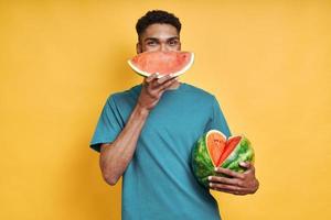 Playful African man holding watermelon while standing against yellow background photo