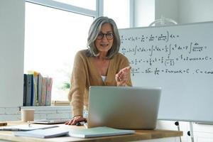 Mature woman teaching mathematics while standing near the whiteboard and looking at laptop photo