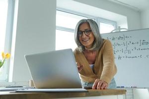 Mature woman teaching mathematics while standing near the whiteboard and looking at laptop photo