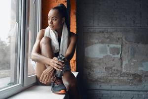 Attractive young African woman holding bottle with water while taking a break in gym