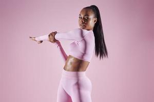 Confident young woman in sports clothing doing stretching exercises against pink background photo