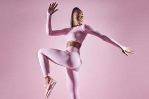 Beautiful young woman in sports clothing exercising against pink background photo