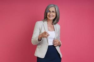 Happy senior woman in formalwear pointing camera while standing against pink background photo