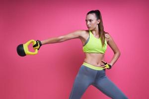 Beautiful young woman exercising with kettlebell against pink background photo