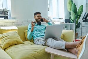 Cheerful African man using laptop and talking on mobile phone while sitting on the couch photo