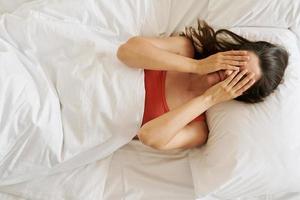 Top view of depressed young woman covering face with hands while lying in bed photo