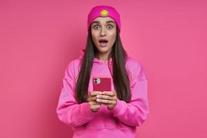 mujer joven sorprendida con un sombrero funky sosteniendo un teléfono inteligente contra un fondo rosa foto